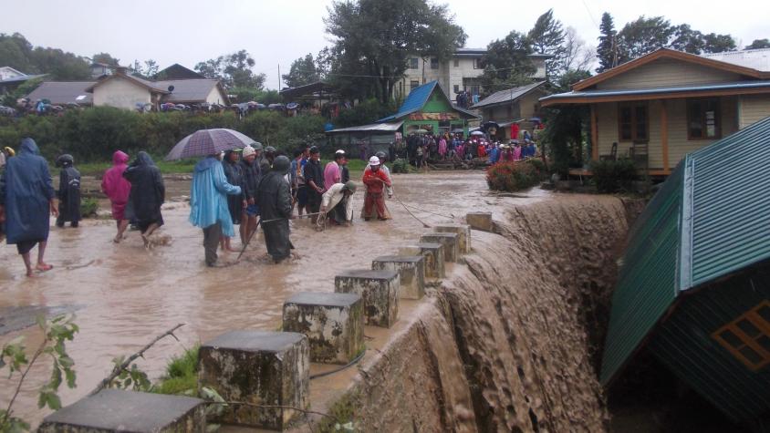 People standing on a flooded road in Myanmar next to a house that washed away.